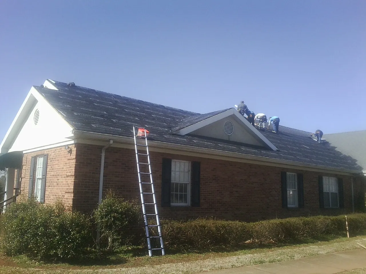 Crew tearing shingles off a brick ranch roof with a ladder set — Emergency Roof Repair work by our crew in Nineveh