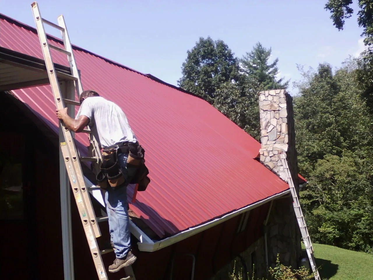 Roofer on a ladder beside a red metal roof and stone chimney — Soffit & Fascia Repair by Roofing Brothers in Nineveh